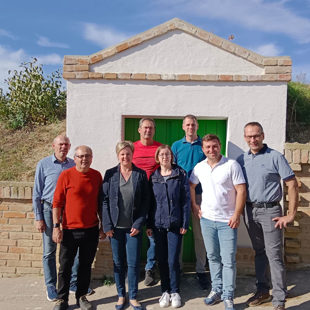Eine Gruppe von sieben Erwachsenen steht vor einem kleinen Gebäude mit einer grünen Tür und Ziegelwänden. Der Himmel ist klar mit verstreuten Wolken.