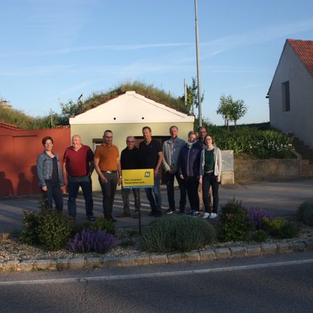 Eine Gruppe von Menschen steht vor einem kleinen Gebäude. Es gibt ein gelbes Schild mit Text. Sie tragen Freizeitkleidung. Das Gebäude hat ein rotes Dach.