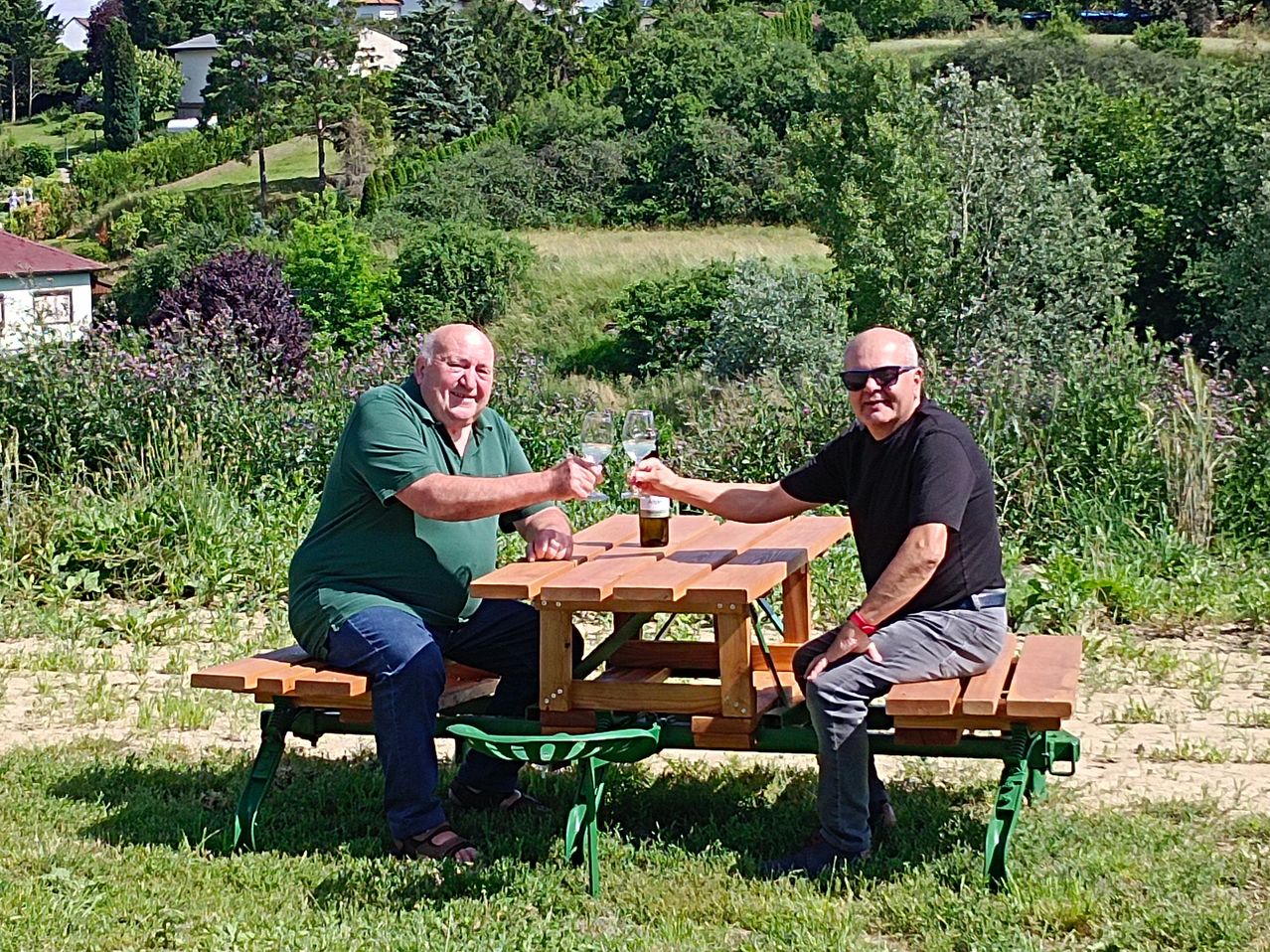 Two men sit at a wooden picnic table in a grassy area, holding wine glasses and smiling. Behind them is a hill with greenery and a house.