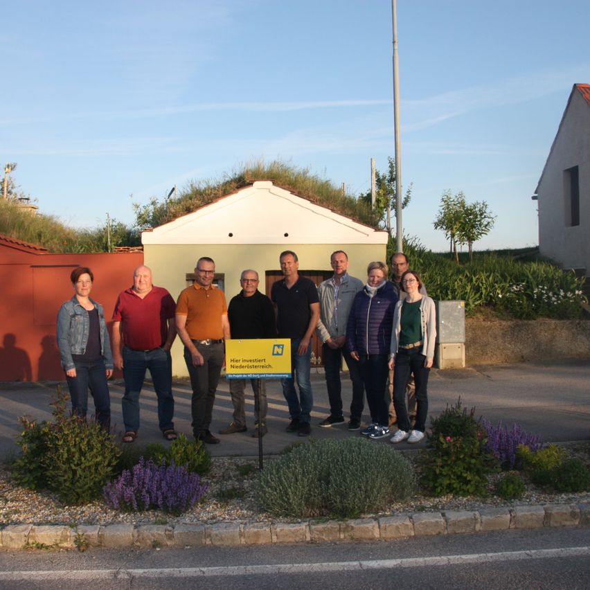 A group of people stands in front of a small house with a yellow sign in front of it. There are plants and flowers on the side of the road.