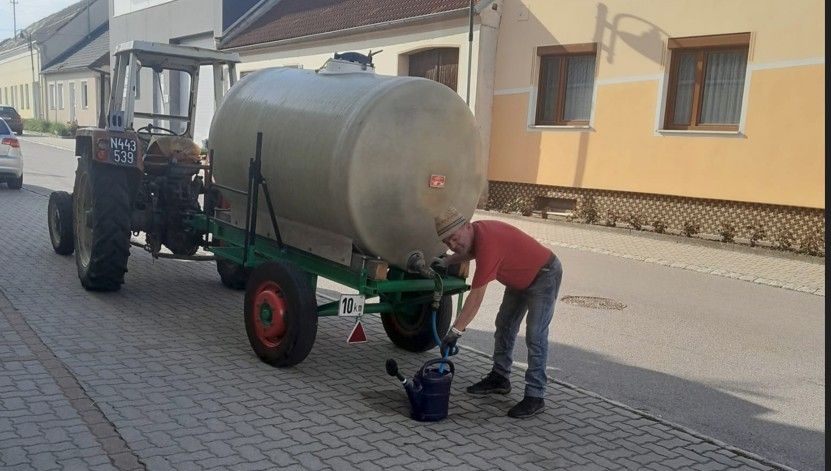 A man wearing a hat and gloves is using a tractor to fill a large water tank with a blue watering can.