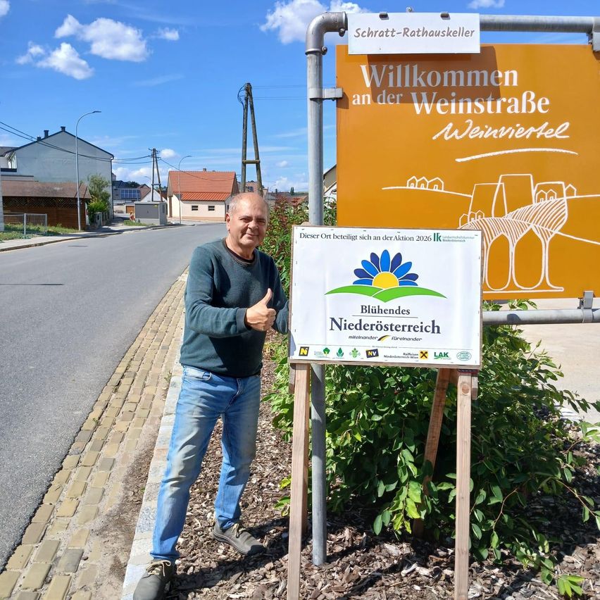 A man stands next to a sign that reads Welcome to the Wine Road Wienersel, and a larger sign with a yellow background and white text.