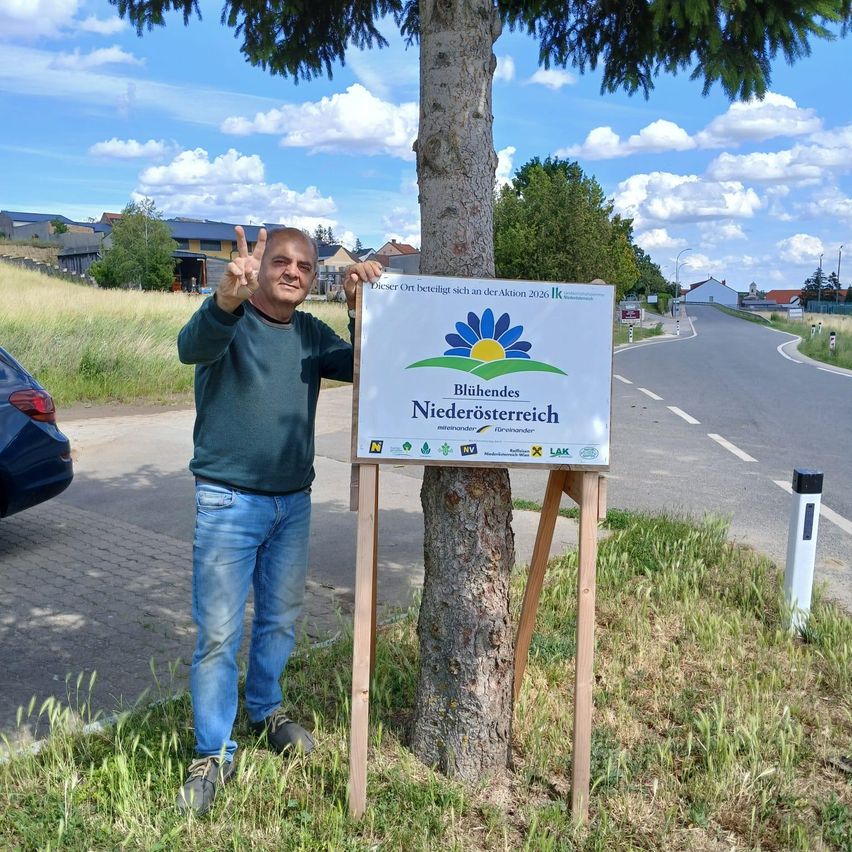 A man stands next to a tree with a sign for the 'Blühende Niederosterreich' action. He makes a peace sign. The sign features a flower and text in German. The scene is outdoors on a sunny day.