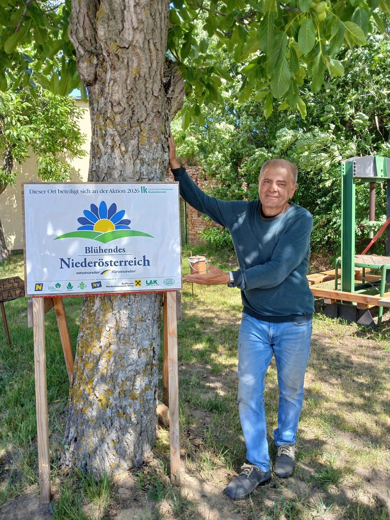 A man stands next to a tree with a sign that reads 'Bluihendes Niederosterreich'. He holds a cup in his hand. Behind him, there is a green structure and benches.
