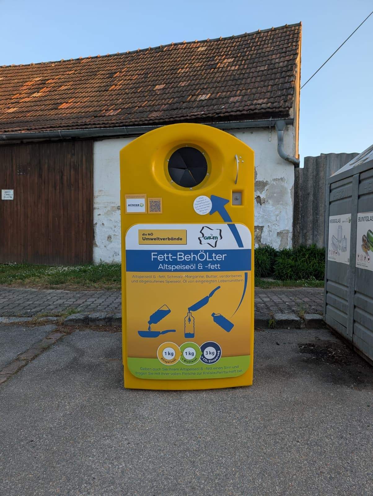 A yellow container for used cooking oil and fat disposal stands in front of an old building. The container is marked with a blue arrow and the words 'Fett-Behälter Altspeiseöl & -fett'.