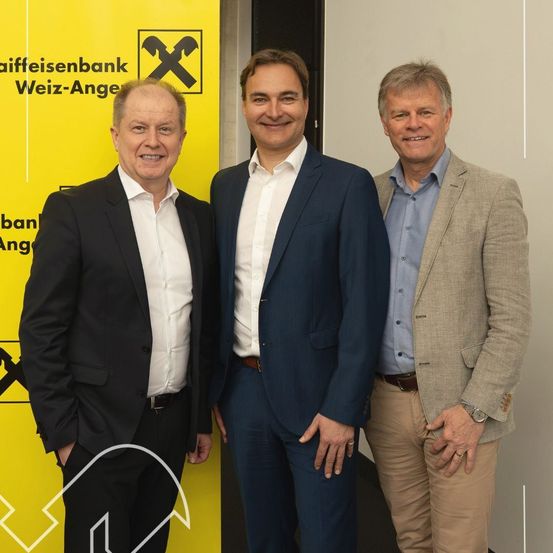 Drei Männer stehen lächelnd nebeneinander vor einem gelben Banner mit der Aufschrift Raiffeisenbank Weiz-Angel.