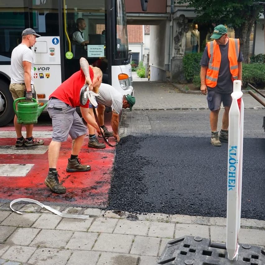 Arbeiter legen neuen Asphalt auf einem Zebrastreifen. Ein Mann hält eine grüne Gießkanne. Ein Bus ist in der Nähe geparkt.
