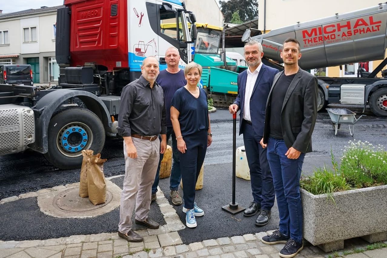 Fünf Personen stehen lächelnd vor Baufahrzeugen. Dahinter befinden sich verschiedene Lastwagen und Maschinen, darunter einer mit der Aufschrift 'MITHER'.