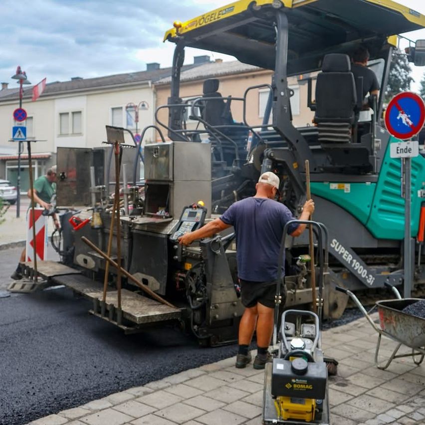 Ein Bauarbeiter lenkt eine große Asphaltiermaschine, gekennzeichnet mit SUPER 1900-3i, während ein anderer Arbeiter die Straße fegt. Sie befinden sich in einer Stadt mit Gebäuden, Fahnen und Verkehrszeichen.