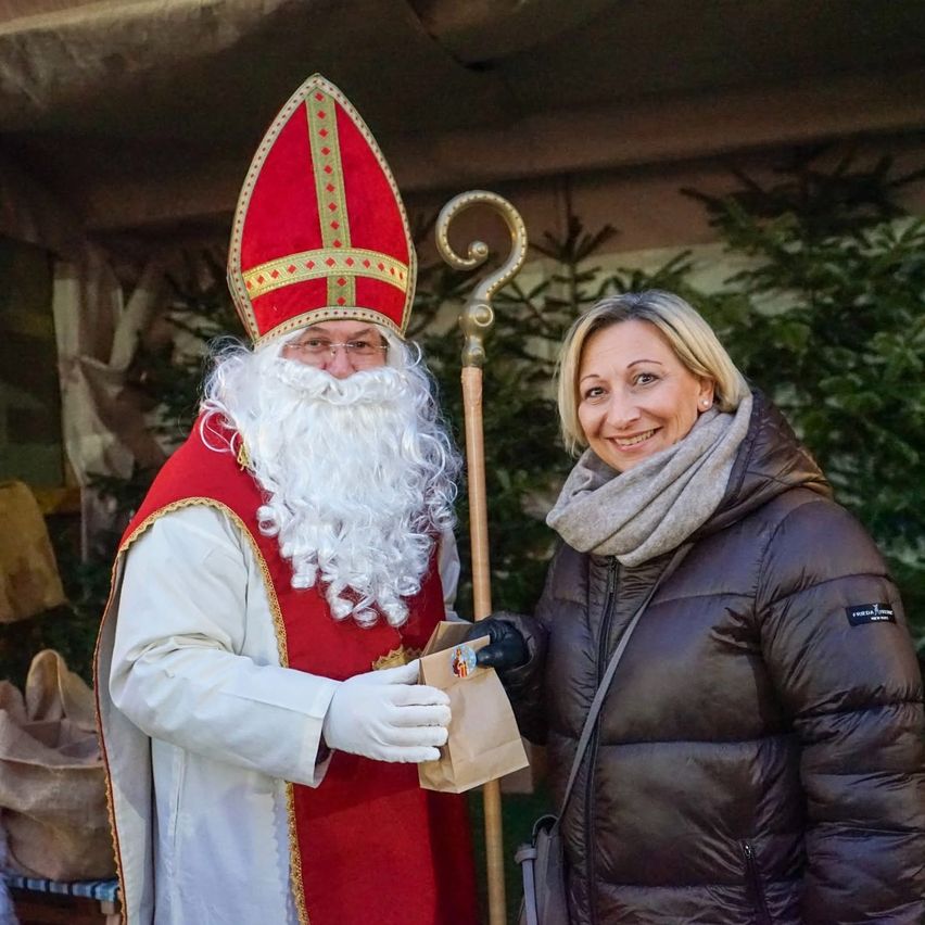 Ein Mann in Santa-Claus-Kostüm hält einen braunen Papierbeutel und lächelt. Neben ihm steht eine Frau mit Schal und Jacke. Sie stehen unter einem Baldachin.