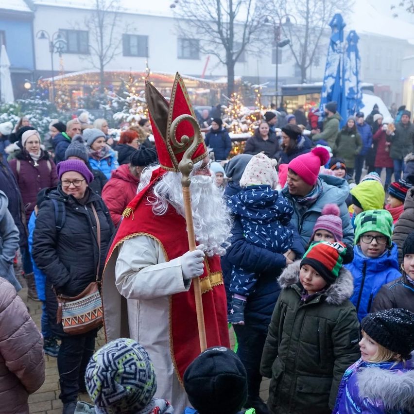 Eine Menschenmenge, bestehend aus Kindern und Erwachsenen, versammelt sich im Freien, wahrscheinlich auf einem Fest. Ein Mann, der als Weihnachtsmann verkleidet ist, interagiert mit der Menge, umgeben von anderen Menschen, einige tragen Mützen und Brillen. Im Hintergrund befinden sich Bäume, Gebäude und Straßenlaternen.