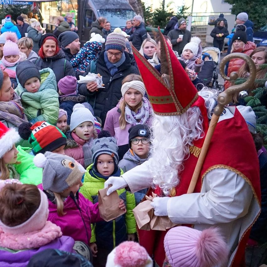 Eine große Menschenmenge, hauptsächlich Kinder, versammelt sich um einen als Weihnachtsmann verkleideten Mann. Er verteilt Papiertüten an die Kinder. Die Menschen sind in Winterkleidung und Mützen gekleidet. Der Schauplatz scheint ein Straßen- oder öffentlicher Raum zu sein.