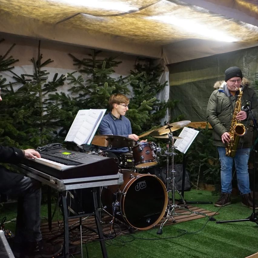 Eine Band spielt Musik in einem Zelt mit Weihnachtsbäumen im Hintergrund. Ein Mann spielt die Saxophon, ein anderer spielt die Drums, und ein dritter spielt das Klavier.