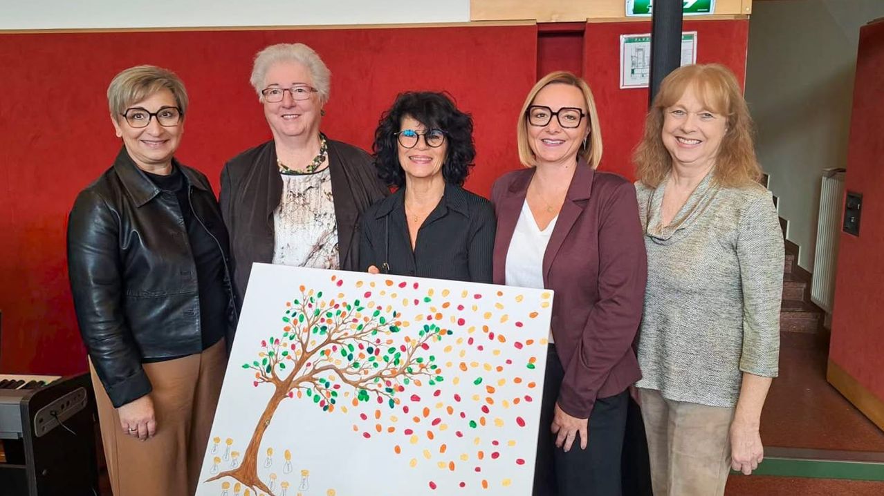 Fünf Frauen stehen hinter einer großen Leinwand mit einem Baum-Zeichnung und lächeln für ein Foto. Sie tragen alle eine Brille. Die Frau in der Mitte hält die Leinwand. Die Wand hinter ihnen ist rot.