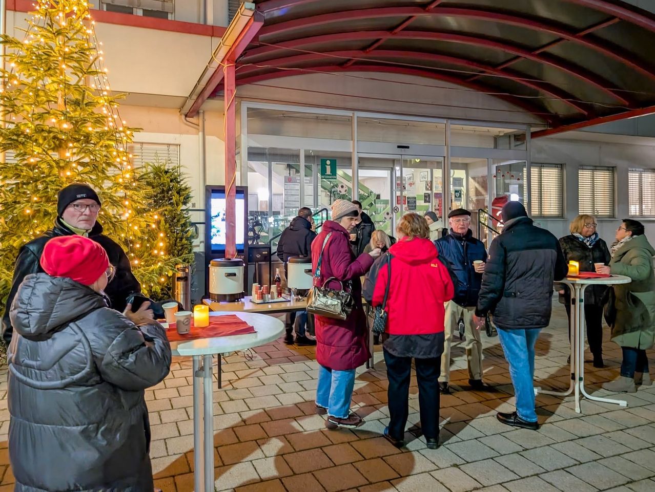 Eine Gruppe von Menschen steht draußen in der Nähe eines Tisches mit einer brennenden Kerze. Sie sind in Winterkleidung gekleidet. Ein beleuchteter Weihnachtsbaum und ein Gebäude mit Glastüren sind im Hintergrund zu sehen.