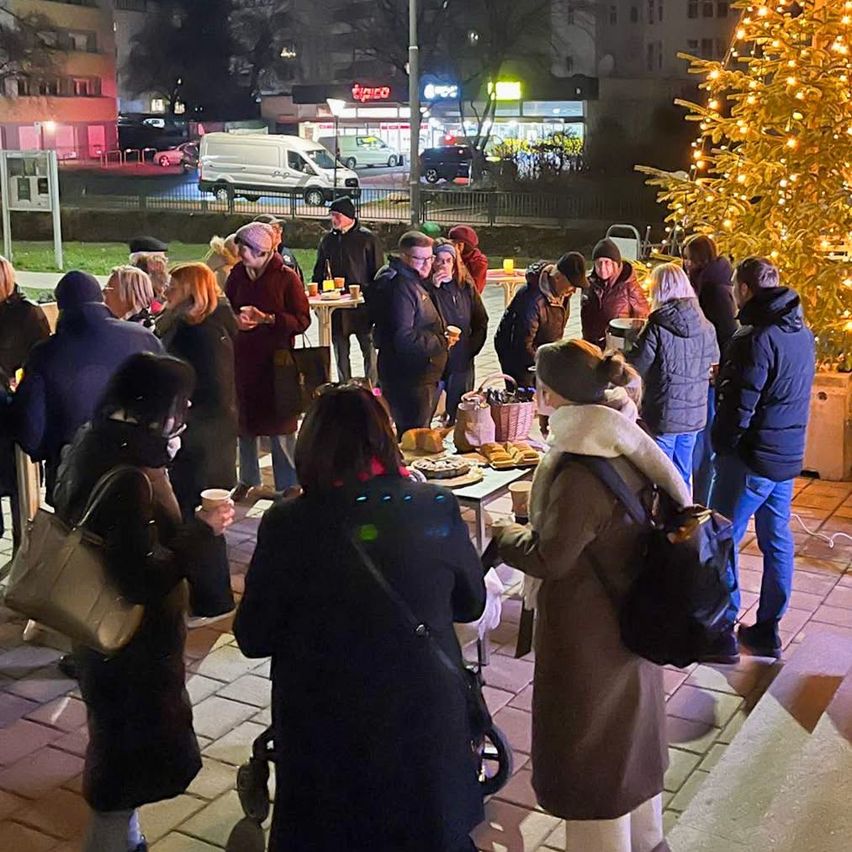Eine Gruppe von Menschen versammelt sich um einen festlich geschmückten Weihnachtsbaum und genießt Essen und Getränke. Die Szene spielt bei Nacht, mit Gebäuden und geparkten Autos im Hintergrund.