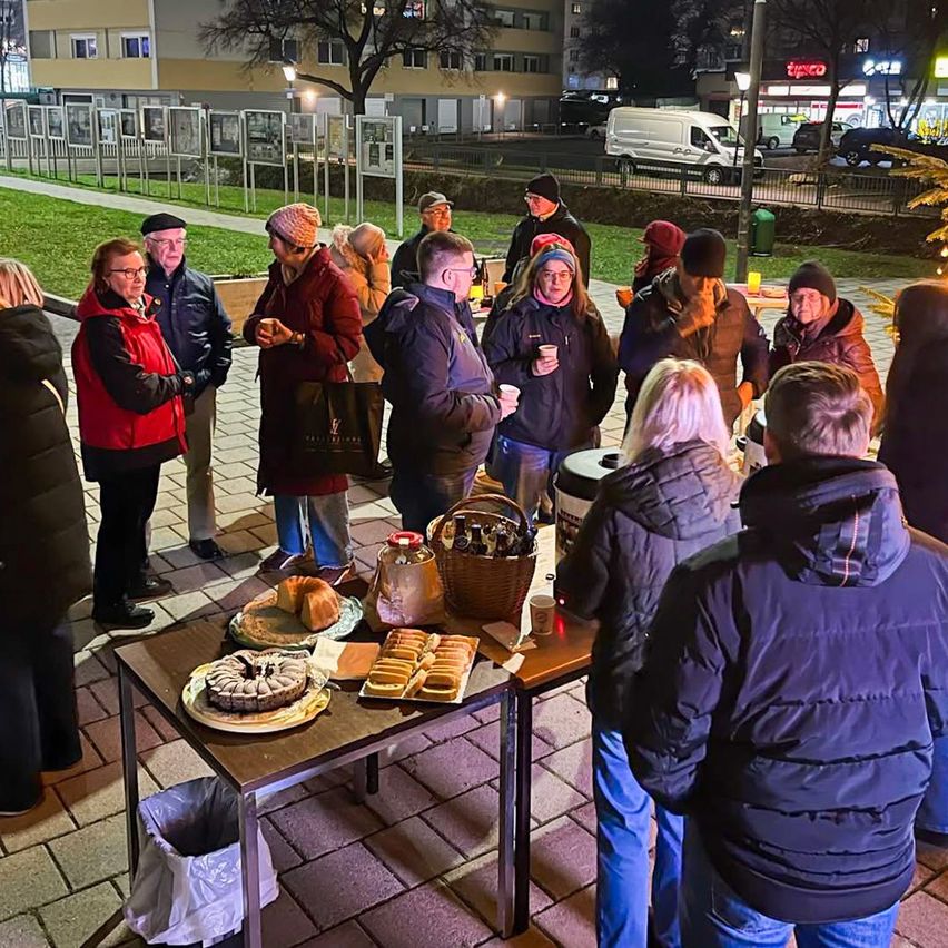 Eine Gruppe von Menschen versammelt sich bei Nacht im Freien, steht um einen Tisch mit Essen, in der Nähe von Gebäuden und geparkten Autos, mit einer festlichen Atmosphäre.