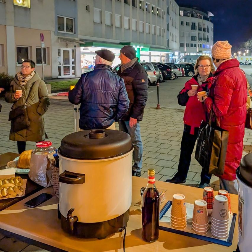 Mehrere Menschen stehen an einem kühlen Abend im Freien und unterhalten sich in der Nähe eines Tisches mit Kaffeemaschine, Brot und Tassen. Im Hintergrund sind Gebäude und geparkte Autos zu sehen.