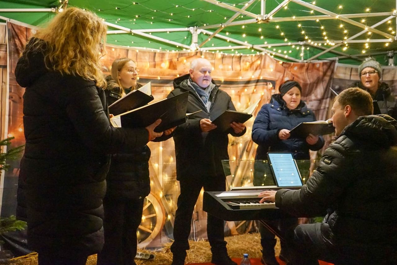 Eine Gruppe Erwachsener versammelt sich unter einem Zelt und singt, während sie Musikbücher halten. Ein Mann spielt Klavier, vor ihm steht ein Laptop auf einem Ständer. Das Zelt ist mit Lichterketten und einem Hintergrund mit einem Rad dekoriert.