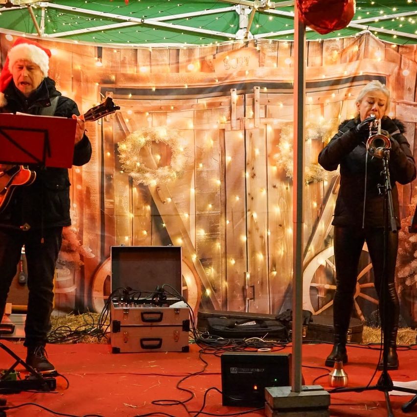 Zwei Personen treten auf einer Bühne mit einem weihnachtlich dekorierten Hintergrund auf. Eine Person spielt Gitarre, während die andere Trompete spielt. Vor ihnen stehen Geräte und ein Mikrofonstativ.