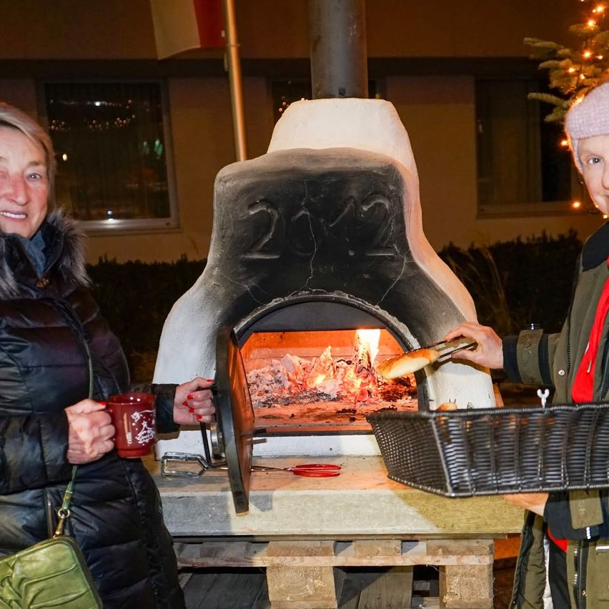 Zwei Frauen stehen neben einem großen Ofen mit Feuer darin. Eine hält einen Korb mit Brot, während die andere einen Becher hält. Sie sind vor einem Gebäude mit einem Weihnachtsbaum im Hintergrund.