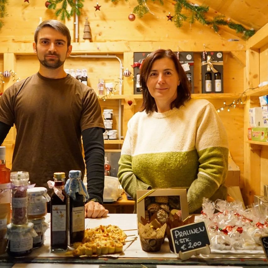Zwei Personen stehen hinter einer Theke in einem rustikalen Laden. Ein Mann mit Bart und eine Frau in einem gestreiften Pullover. Sie haben verschiedene Lebensmittel zur Schau gestellt.