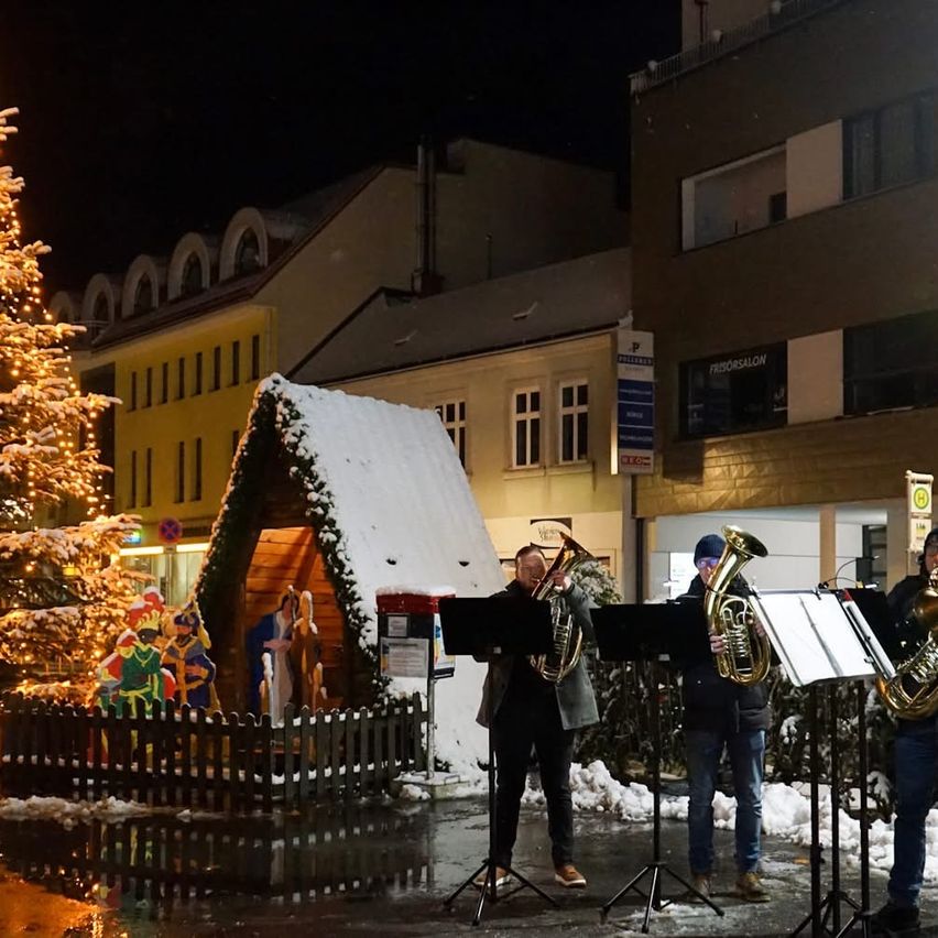 Drei Musiker spielen Instrumente vor einem Weihnachtsbaum auf einem Stadtplatz. Dahinter steht eine Krippe. Gebäude umgeben das Gebiet.