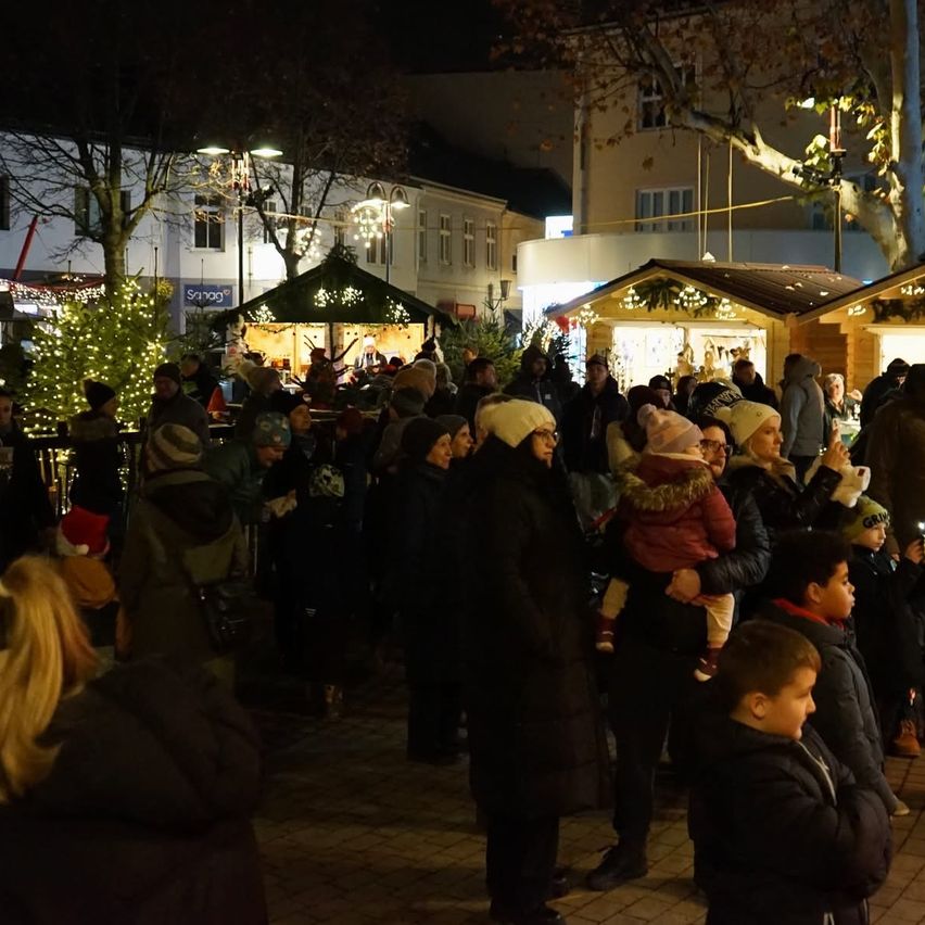 Ein überfüllter Außenbereich bei Nacht mit vielen Menschen, einschließlich Kindern, die sich um einen geschmückten Weihnachtsbaum und festliche Stände versammelt haben.