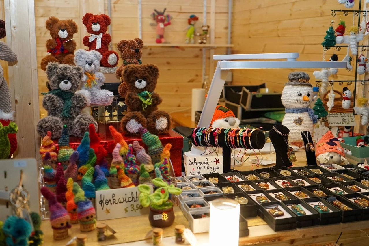 Eine Ausstellung von gestrickten Spielzeugen umfasst Bären, Gnome und einen Schneemann. Verschiedene Accessoires sind in Regalen zu sehen, einschließlich Armbänder, Halsketten und Ringe. Das Preisschild zeigt 3,50 für die Gnome.