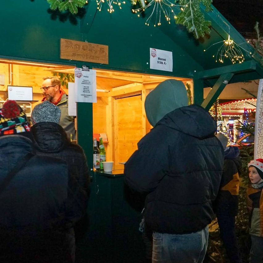 Menschen stehen in einer Schlange an einem Stand mit grünem Dach, beleuchtet von Lichterketten. Eine Person im Inneren bedient sie. Der Stand hat ein Schild mit der Aufschrift 'Maroni 9.30k.4'.