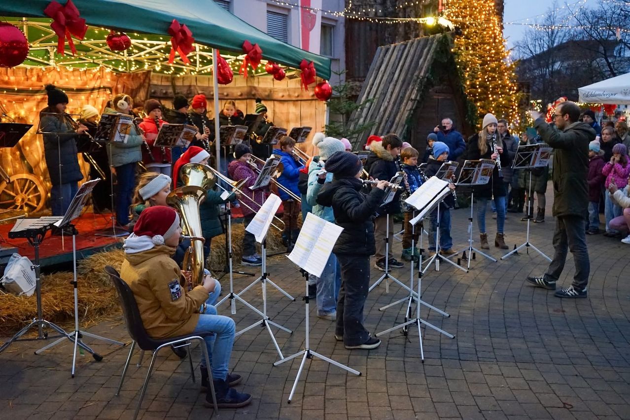 Eine Gruppe von Musikern spielt Instrumente unter einem Zelt mit Weihnachtsdekoration. Sie tragen Winterkleidung und Mützen, mit Zuschauern hinter ihnen. Die Umgebung umfasst einen festlich beleuchteten Baum und ein Gebäude.