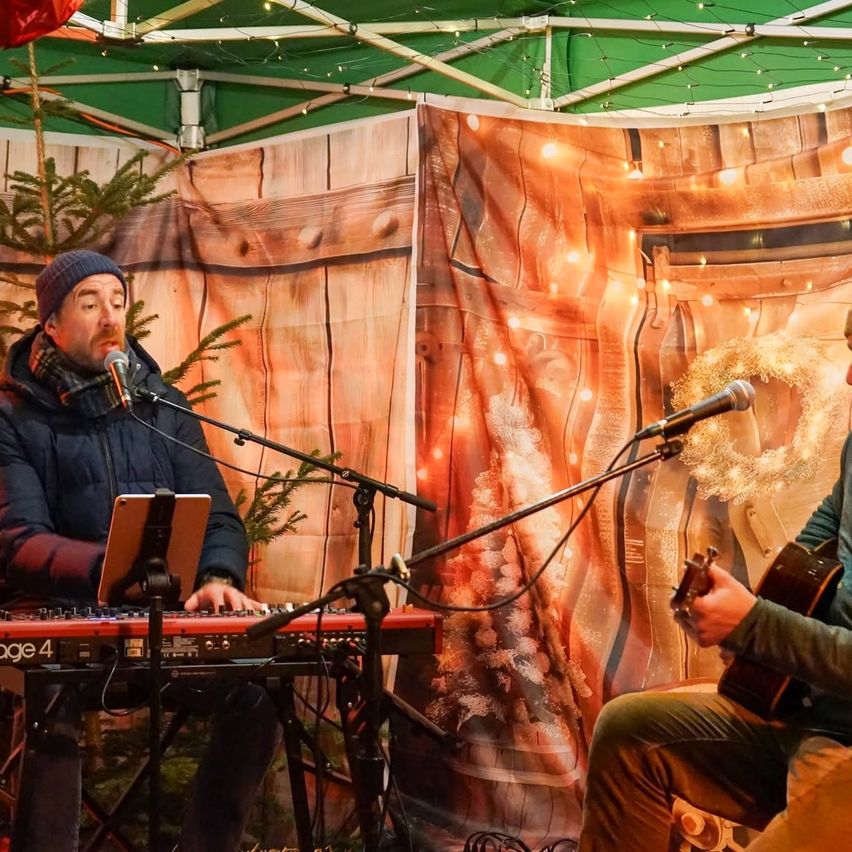 Zwei Männer spielen Musik. Einer spielt die Tastatur und der andere die Gitarre. Sie sind unter einem grünen Zelt mit Holzdekorationen und Lichtern.