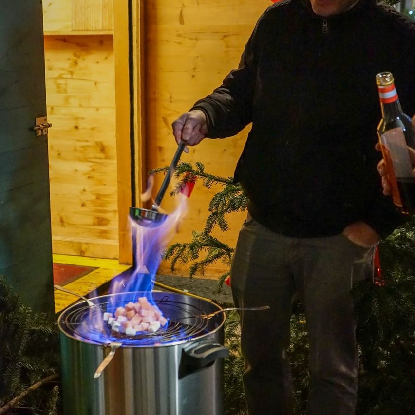 Ein Mann kocht Marshmallows über einer blauen Flamme aus einem Topf auf einem Feuer, hält eine Schöpfkelle und eine Flasche. Der Hintergrund ist innen mit einer Holzwand und Weihnachtsdekoration.