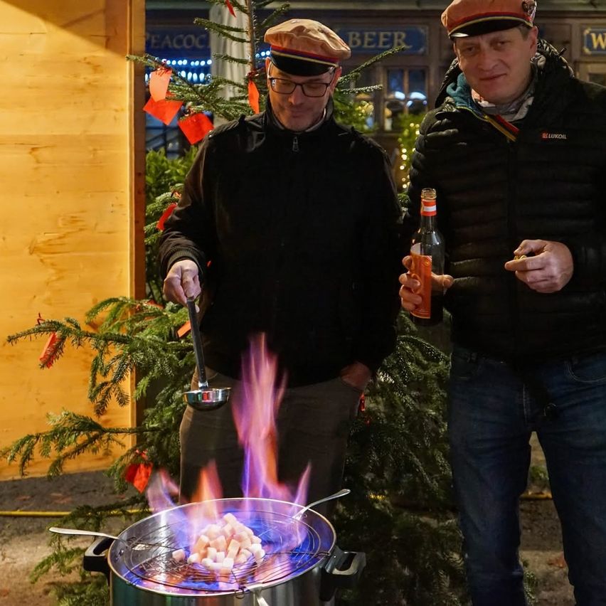 Zwei Männer kochen im Freien mit einem Grill über offenem Feuer vor einem geschmückten Weihnachtsbaum. Ein Mann hält einen Schöpflöffel und eine Flasche, der andere eine Zigarette. Dahinter steht ein Gebäude mit einem Bierschild.