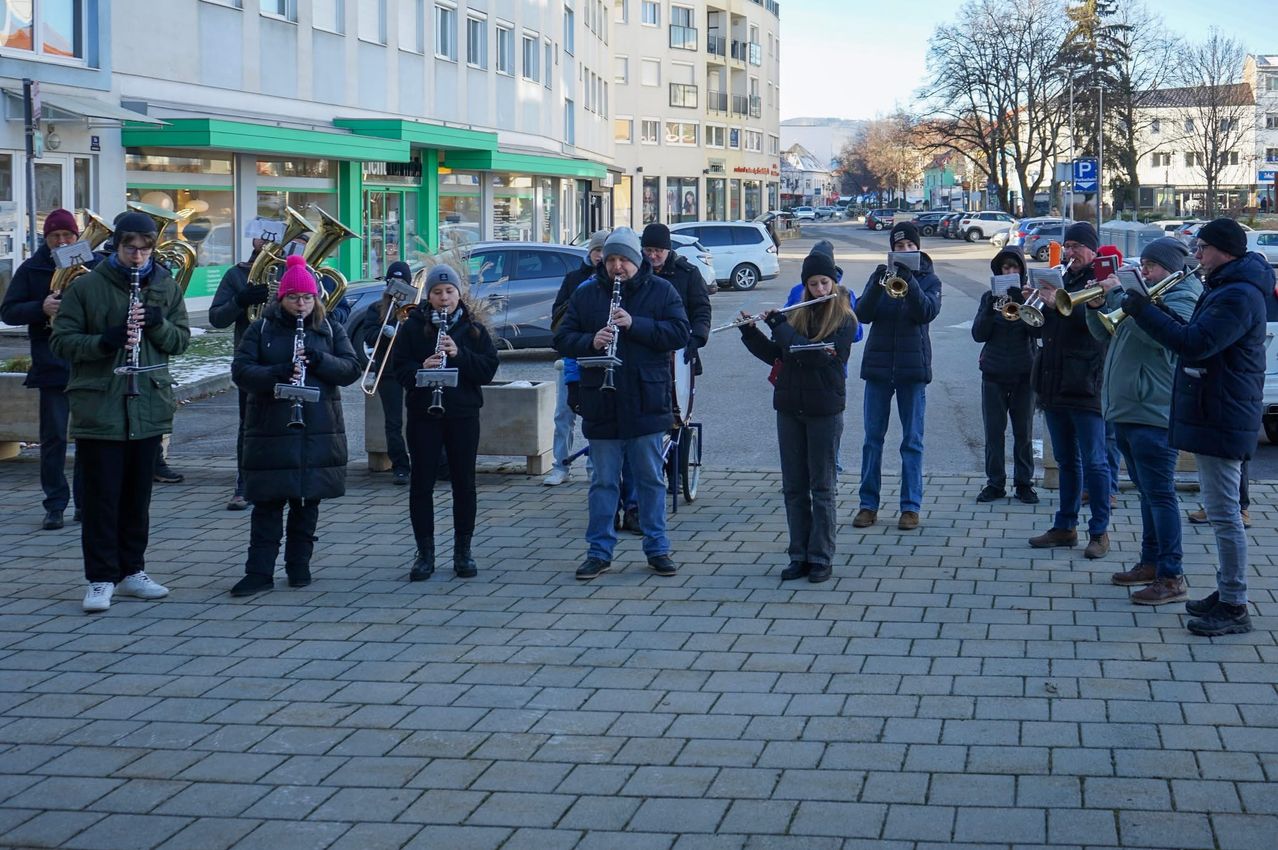 Eine Gruppe von Menschen in Winterkleidung spielt Musikinstrumente auf einer gepflasterten Straße in einer Stadt. Im Hintergrund sind Gebäude und geparkte Autos zu sehen.