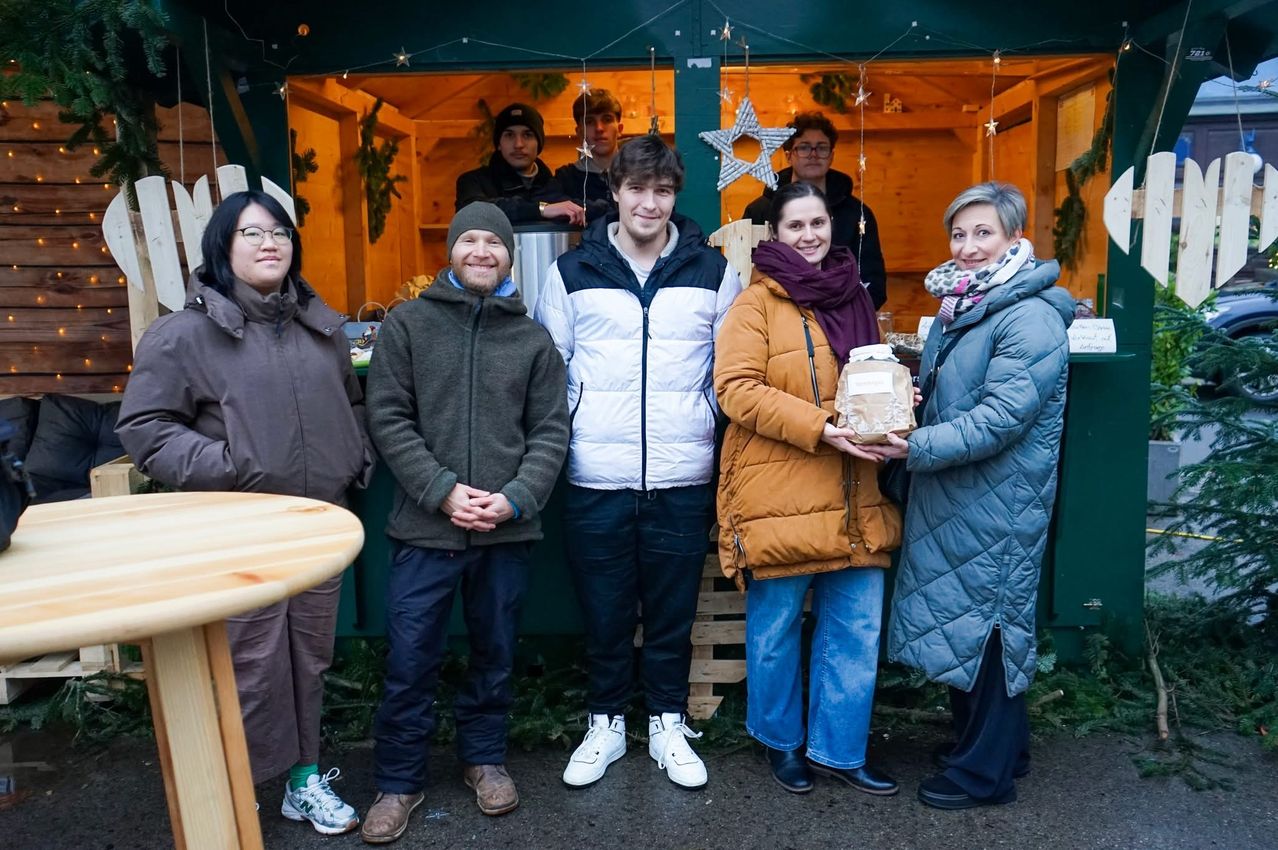 Eine Gruppe von fünf Personen steht lächelnd vor einem dekorierten Stand. Eine Frau hält ein Paket. Dahinter steht ein Mann, der einen Topf bedient, und ein weiterer steht im Inneren. Die Umgebung wirkt festlich.