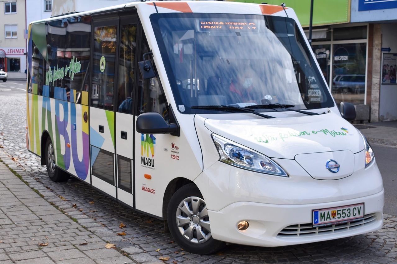 Ein weißer Bus mit bunten Designs an der Seite, geparkt auf einer gepflasterten Straße. Die Front hat ein Nummernschild mit 'MA'. Das Fahrerfenster zeigt Reflexionen von nahen Gebäuden.