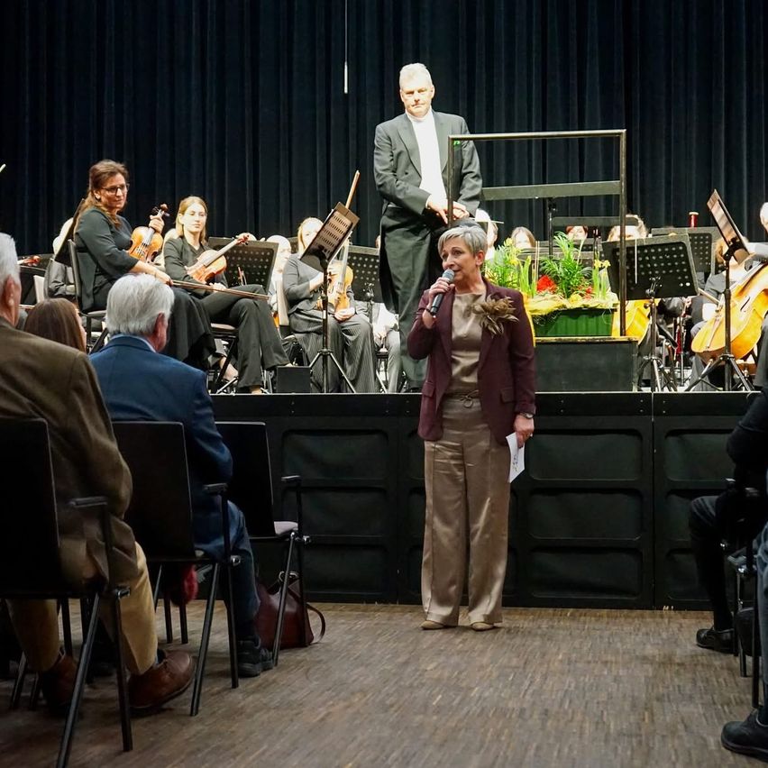 Eine Frau auf der Bühne spricht in ein Mikrofon vor einem Orchester und einem Publikum. Hinter ihr steht ein Mann mit den Händen im Rücken.