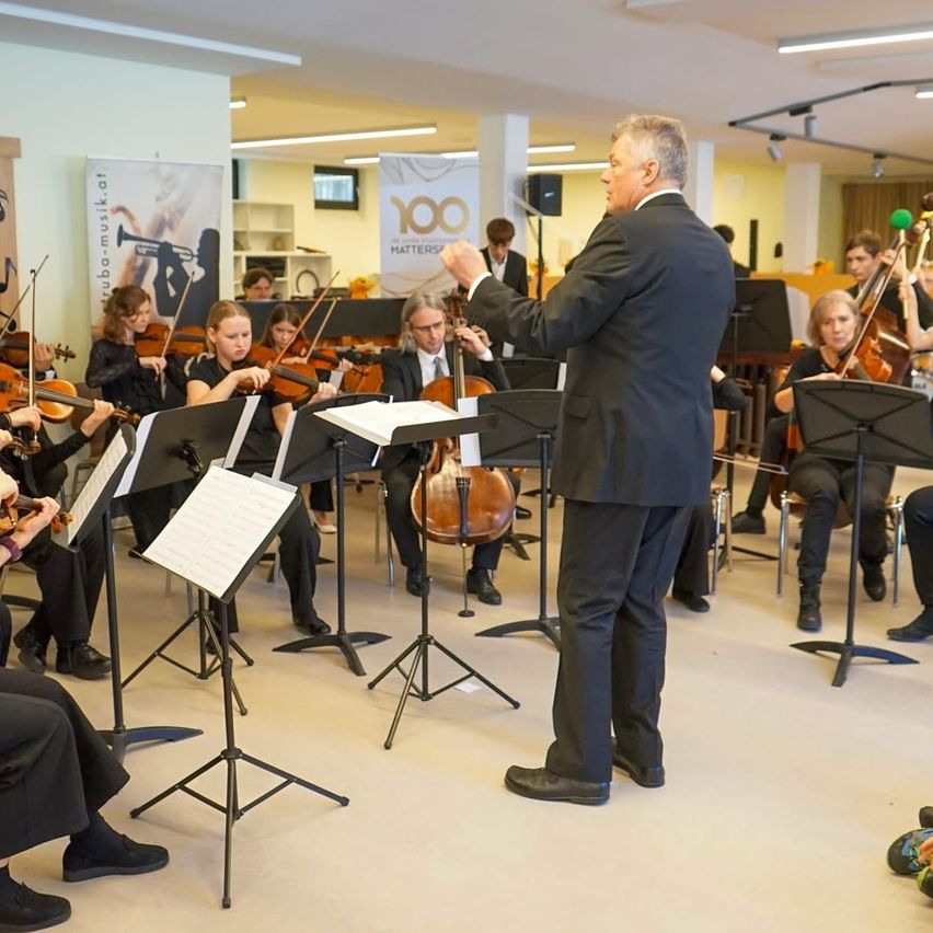 Ein Dirigent führt ein Streichorchester in einem Raum an, mit mehreren Musikern, die Violinen und Celli spielen. Zuschauer sitzen davor.