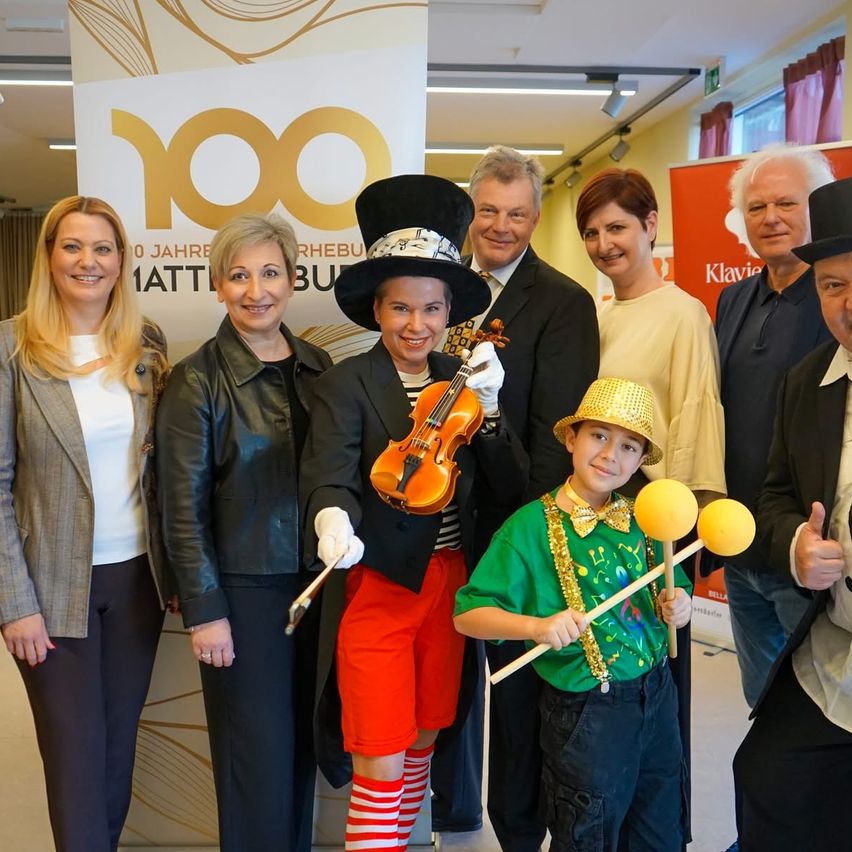 Eine Gruppe von Menschen in Kostümen und Hüten posiert für ein Foto in einem Raum mit einem Schild, das 100 Jahre anzeigt. Eine Frau in einer schwarzen Lederjacke hält eine Geige.