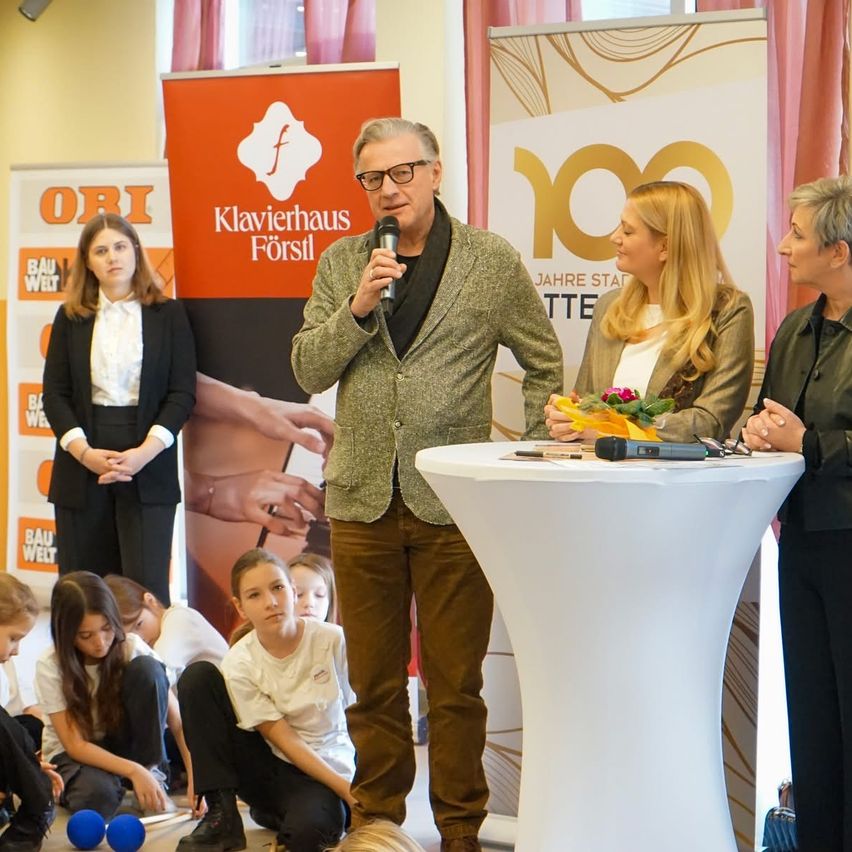 Ein Mann mit Brille spricht in ein Mikrofon am Podium, umgeben von drei Frauen, von denen eine Blumen hält. Hinter ihnen befinden sich zwei Banner, eines mit 'Klavierhaus Försil' und das andere mit '100 Jahre'. Kinder sitzen auf dem Boden, und ein Mädchen schaut den Mann an.