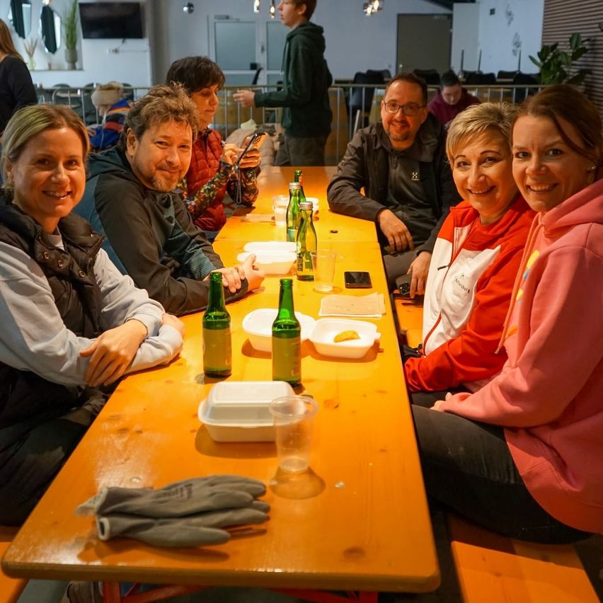 A group of people sits at a wooden table, smiling for a photo. The table has plates, bottles, and glasses. Another person stands behind them, and a TV is mounted on the wall.