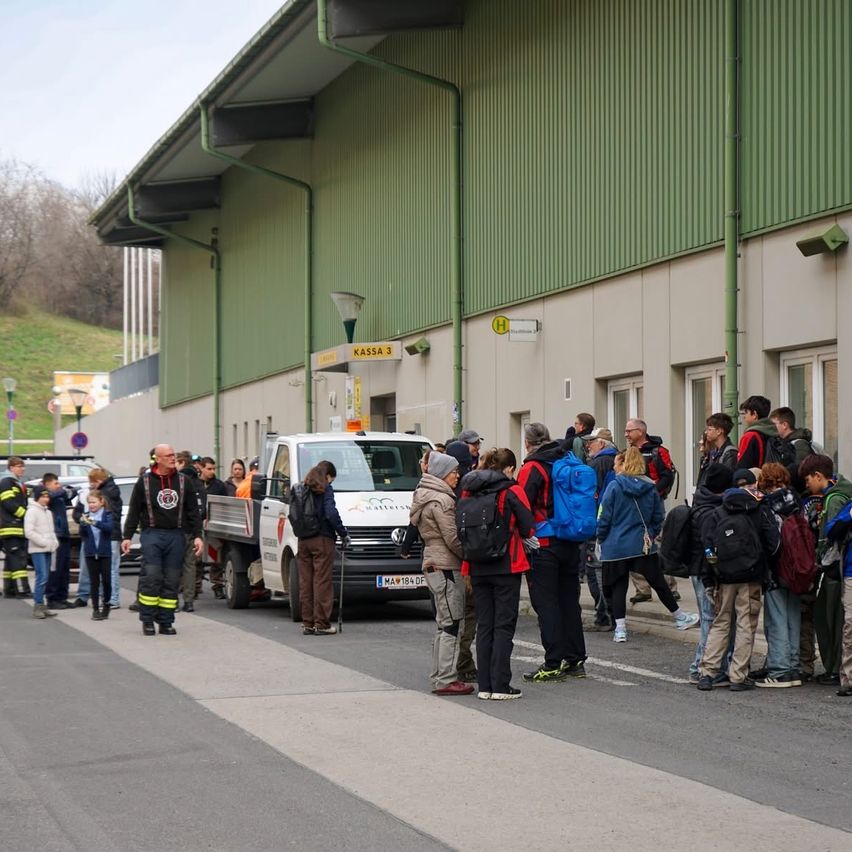 A group of people wait outside a building, with a pickup truck parked nearby. Some individuals wear backpacks and jackets. A person with a cane stands in the middle.