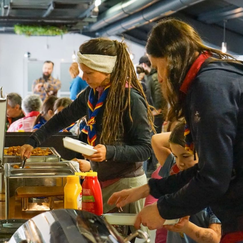 A woman with dreadlocks and a bandana serves food to others at a buffet. Nearby, others eat and a few people stand in the background.