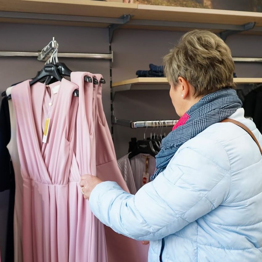 A woman in a winter jacket examines a pink dress in a store, with other clothes on hangers behind her.