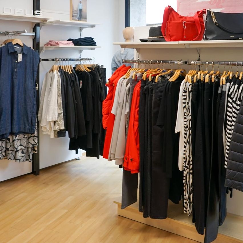 A clothing store displays various outfits on hangers. Shelves hold accessories like bags and hats. A mannequin is in the background.
