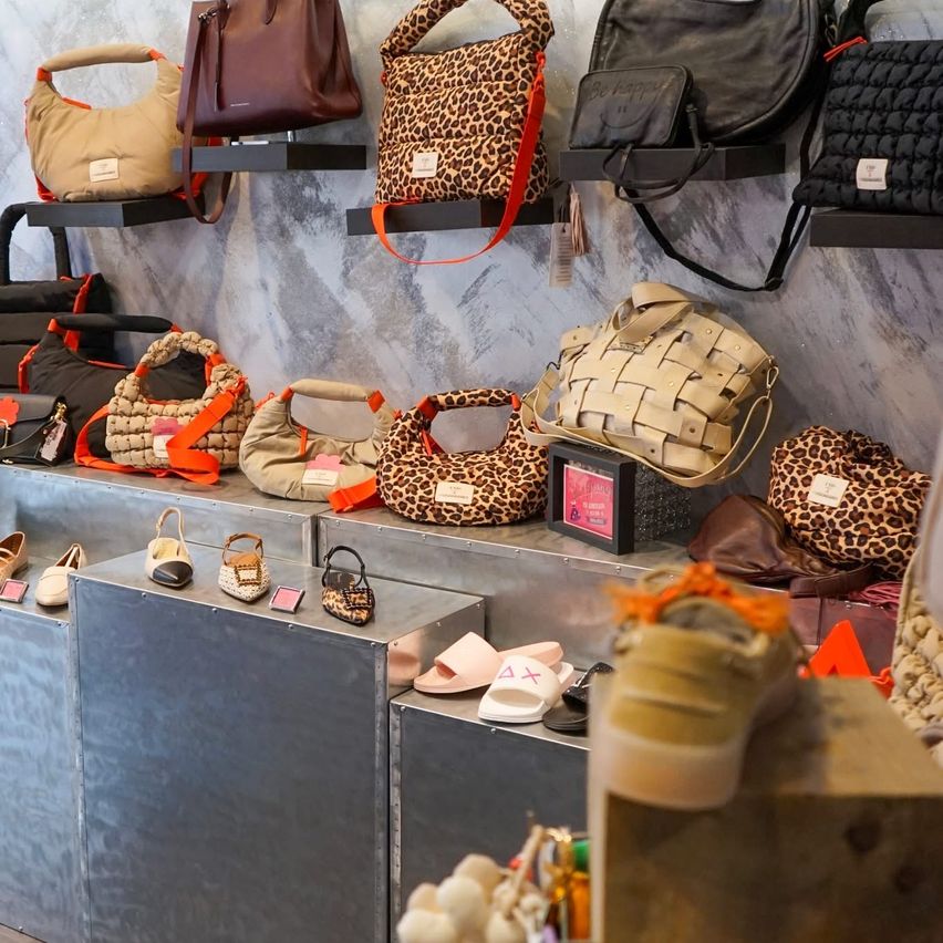 A boutique displays various handbags on shelves and metal racks. Several leopard-print and woven bags hang on black shelves, while others are on display below. Shoes, slippers, and other accessories are also present.