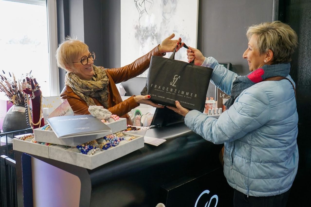 Two women are at a counter, one hands a bag to the other. The bag is black with the text 'FRAUENZIMMER'. Both are smiling.