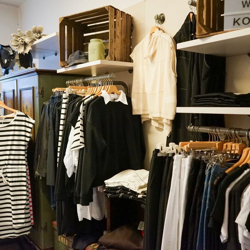 A clothing store with various racks holding different clothing items. Wooden boxes and a shelf with a white sign are on the wall.