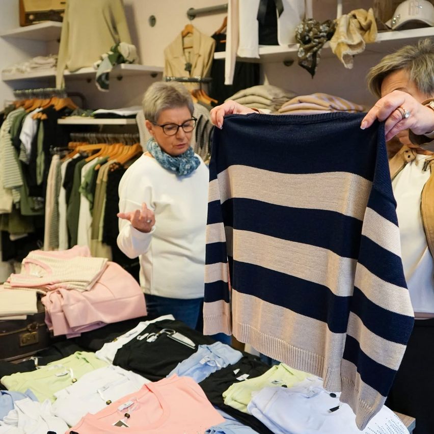 Two women examine a striped sweater in a boutique, surrounded by neatly arranged clothing on shelves and racks.